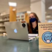 Woman sits out of focus at a table in the library with a mask on and a sign about masks in front of her.
