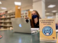 Woman sits out of focus at a table in the library with a mask on and a sign about masks in front of her.