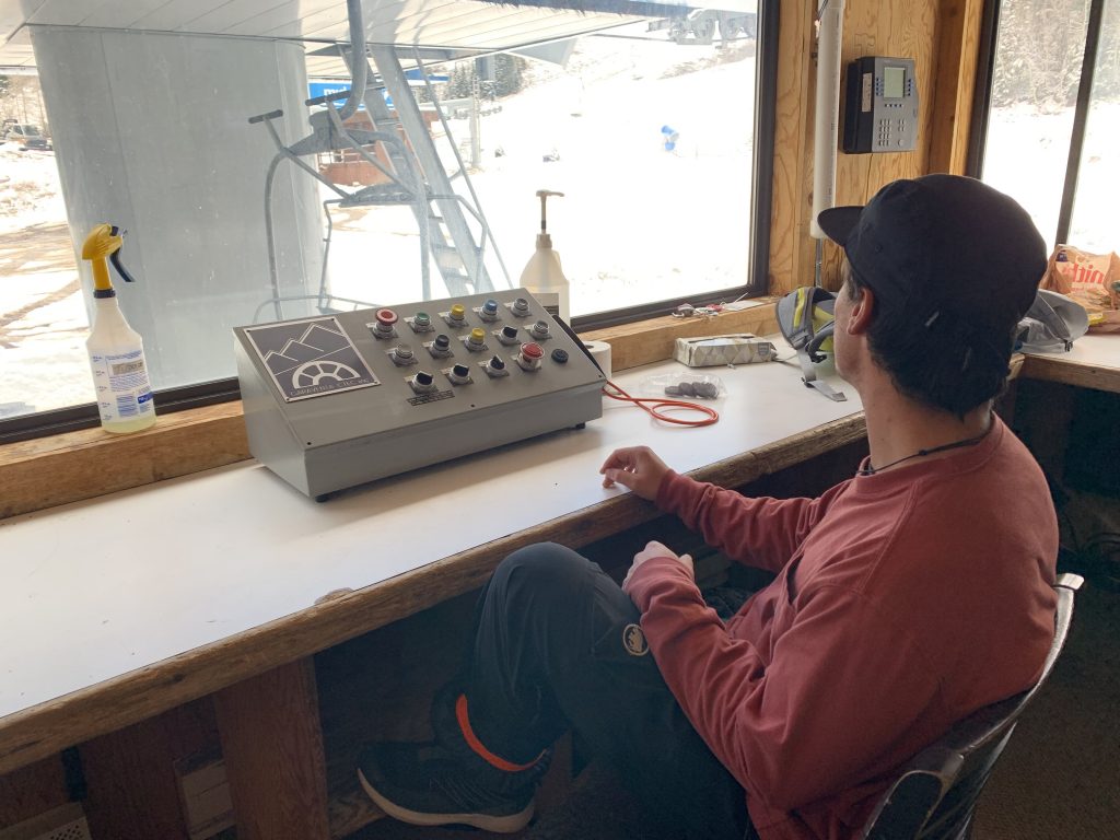 John Brindle sits in a red shirt in an empty room with windows overlooking the snowy hills operating the ski lift with a control panel in front of him. 