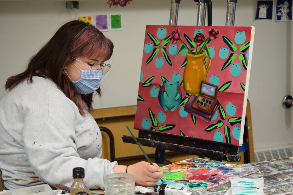 Aunika Woodbry sits next to a painting with a pink background and teal and yellow froge with green plants. She sits next to her painting dipping her brush in the blue pain in a paint pallet covered in shades of green, blue, red, and pink. 