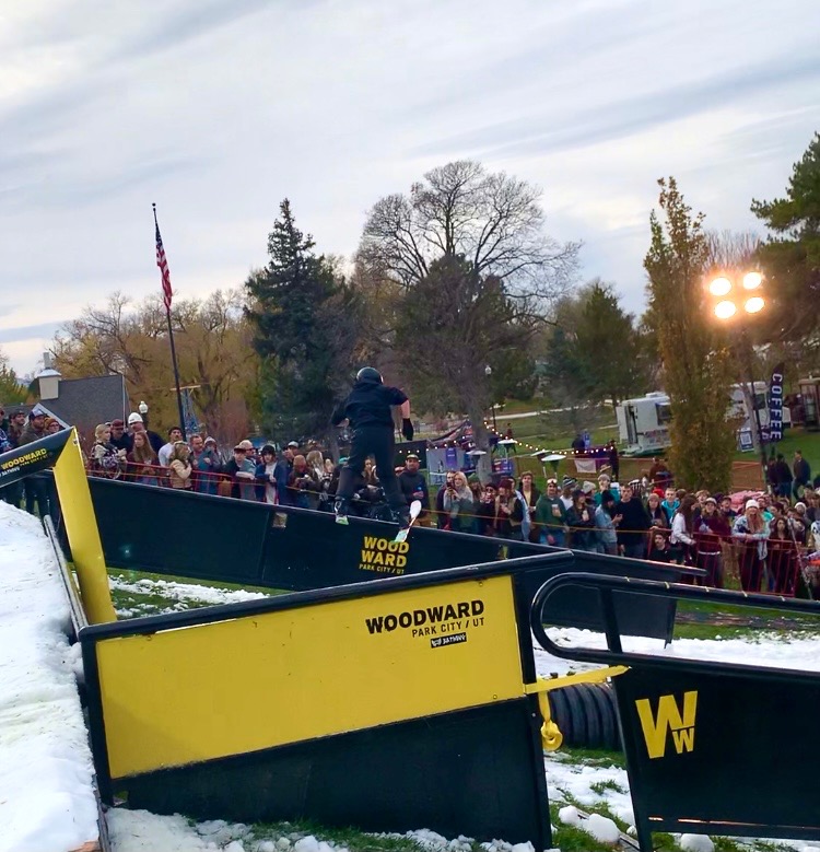 a skiier goes down a black rail on a grass and snow covered hill in front of a crowd watching behind gates. 