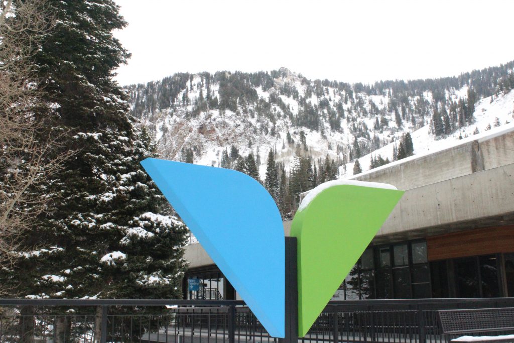 The Snowbird Skii resort logo in blue and greet sits in front of the snowy mountain range with trees in the distance. 