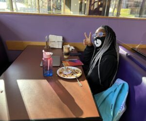 A person sits at a booth in Shaw Student center, smiles at the camera, and makes a peace sign symbol with her right hand.