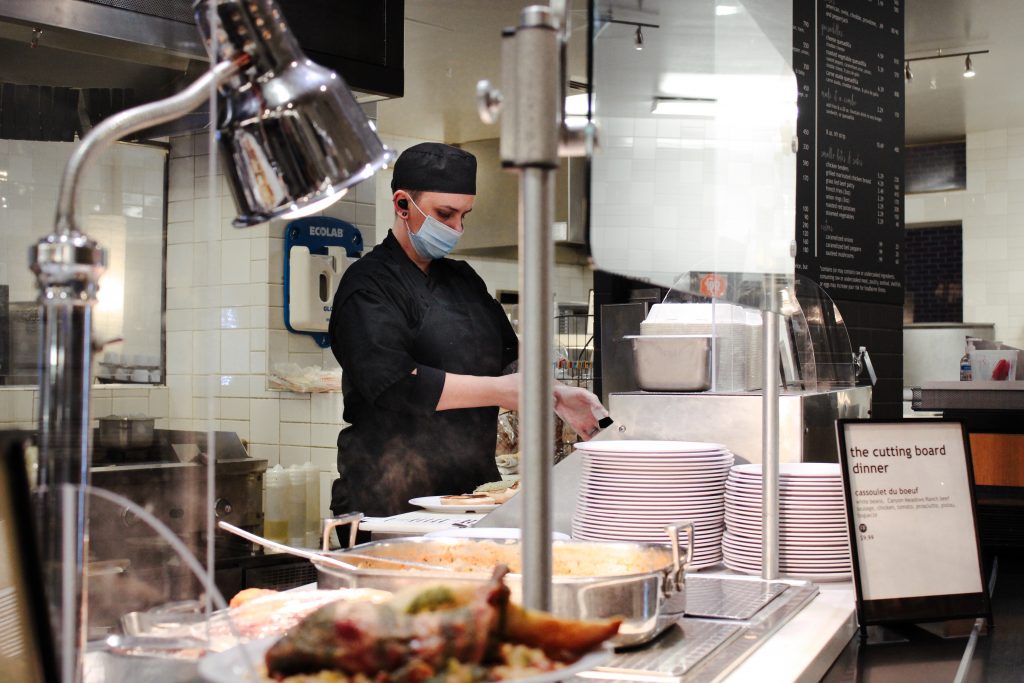A chef puts on food handlers gloves in the middle of the Shaw Cutting Board kitchen amid steaming plates of food.