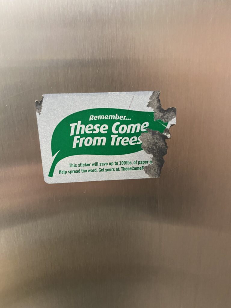 A worn-out sticker on a silver paper towel dispenser with “Remember these come from trees” written on a graphic of a green leaf.