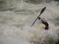 A whitewater kayaker paddles through a large rapid while water crashes over the bow of the kayak.
