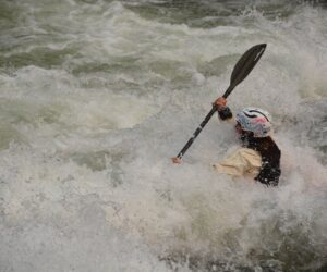 A whitewater kayaker paddles through a large rapid while water crashes over the bow of the kayak.