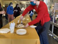 A woman in a red sweater serves chocolate pie at a long banquet table at the Annual Staff Council Pie Auction in Bassis Student Center as patrons gather around.