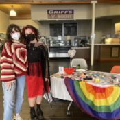 Sophie Caligiuri and Mariah Trujillo with masks stand in front of a table with a rainbow tablecloth and painted rocks.