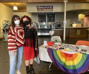 Sophie Caligiuri and Mariah Trujillo with masks stand in front of a table with a rainbow tablecloth and painted rocks.