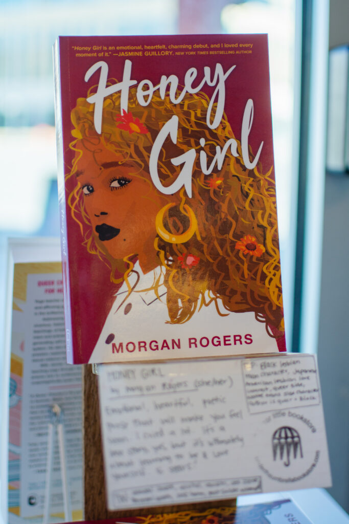 Honey Girl by Morgan Rogers, a new adult fiction novel with a black woman on the cover, sits on a display with a notecard beneath it at Under the Umbrella, a queer bookstore.