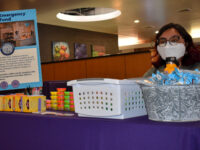 A woman with a mask and glasses sits behind a purple-clothed table with a sign “Student Emergency Support Fund” and other non legible text in the Shaw Student Student March 2. The table is also filled with rice crispy treats, tea boxes, and Play-doh.