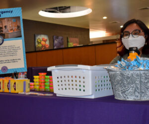A woman with a mask and glasses sits behind a purple-clothed table with a sign “Student Emergency Support Fund” and other non legible text in the Shaw Student Student March 2. The table is also filled with rice crispy treats, tea boxes, and Play-doh.