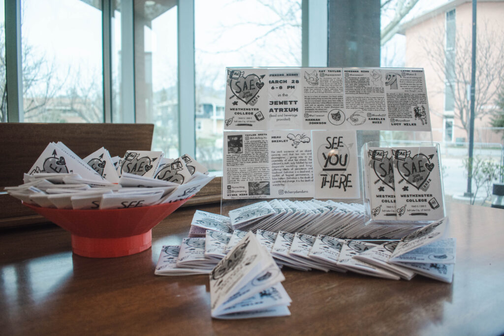 Small white and black zines are propped up, inside an orange bowl, and splayed across a piano’s back in Tanner Atrium in Jewett Center for the Performing Arts on March 28. A few legible words on the zines are SAE, Jewett Atrium and See you there. 