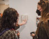 A student with long, wavy black hair and a student with straight blonde hair take a detailed look at an art journal at the Senior Art Exhibit in Tanner Atrium in Jewett Center for the Performing Arts on March 28. The art journal is pinned onto a temporary white wall. Glimpses of more artwork are visible in the background.