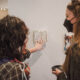 A student with long, wavy black hair and a student with straight blonde hair take a detailed look at an art journal at the Senior Art Exhibit in Tanner Atrium in Jewett Center for the Performing Arts on March 28. The art journal is pinned onto a temporary white wall. Glimpses of more artwork are visible in the background.