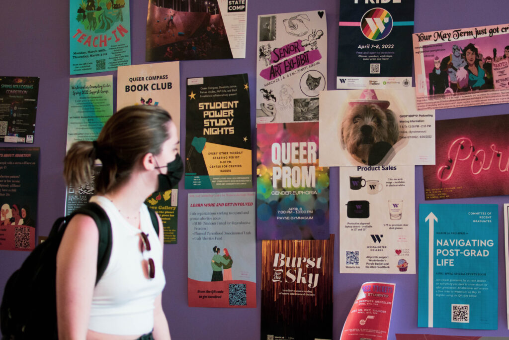 Adia Estes, a white person carrying a backpack, walks past a purple wall covered in posters advertising events on campus. 