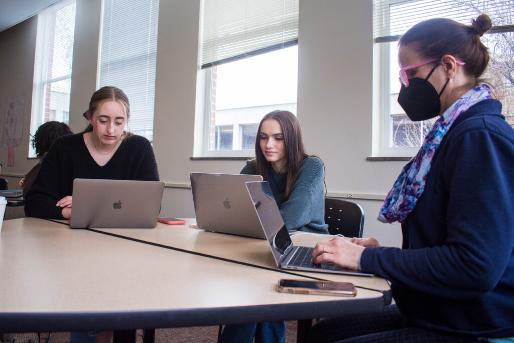 Ashlee Szwedko and Liliana Sauro, both sophomore neuroscience majors, sit without masks at a table with Meghan Wall, a dance professor, who is wearing a mask during class. They are all working on their laptops. 