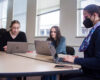Ashlee Szwedko and Liliana Sauro, both sophomore neuroscience majors, sit without masks at a table with Meghan Wall, a dance professor, who is wearing a mask during class. They are all working on their laptops.