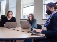 Ashlee Szwedko and Liliana Sauro, both sophomore neuroscience majors, sit without masks at a table with Meghan Wall, a dance professor, who is wearing a mask during class. They are all working on their laptops.