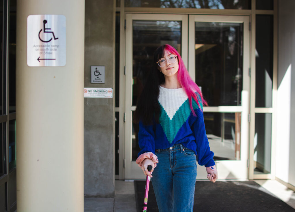 Binnie Green Morris, a white person with long black and pink hair, walks away from a doorway. They are using a pink cane and pass two signs that say the accessible entrance is on the other side of the building. 