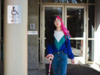 Binnie Green Morris, a white person with long black and pink hair, walks away from a doorway. They are using a pink cane and pass two signs that say the accessible entrance is on the other side of the building.