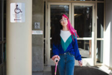 Binnie Green Morris, a white person with long black and pink hair, walks away from a doorway. They are using a pink cane and pass two signs that say the accessible entrance is on the other side of the building.
