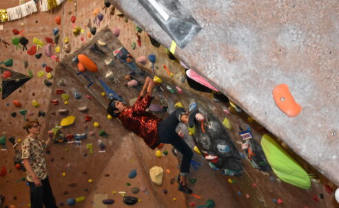 Gabe Medina, a male student climbs a climbing wall, as Travis Goodin, a male student, watches Medina.