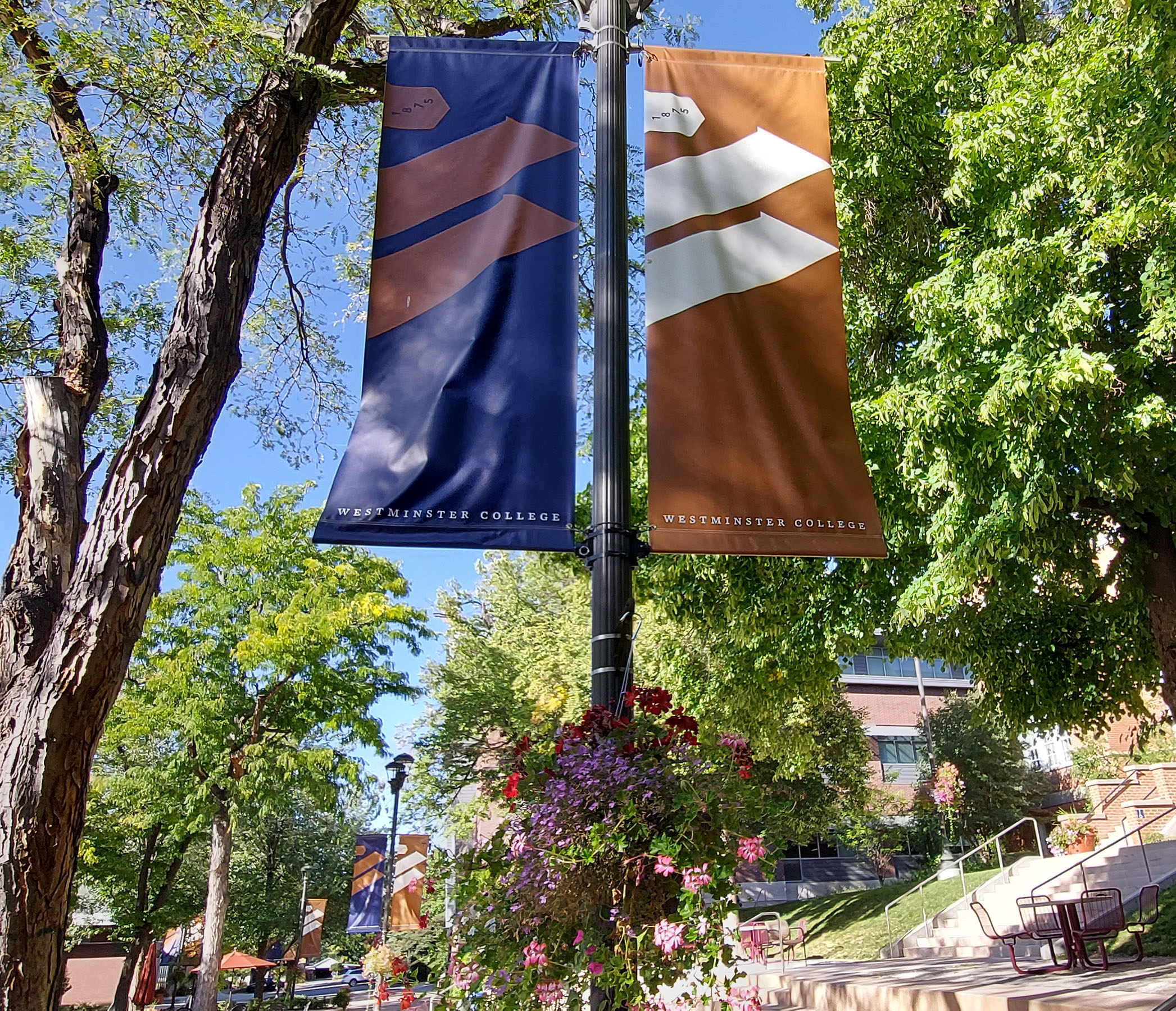 Two Westminster College banners, copper and dark purple, and a flower basket hang from a light post in Richer Commons.