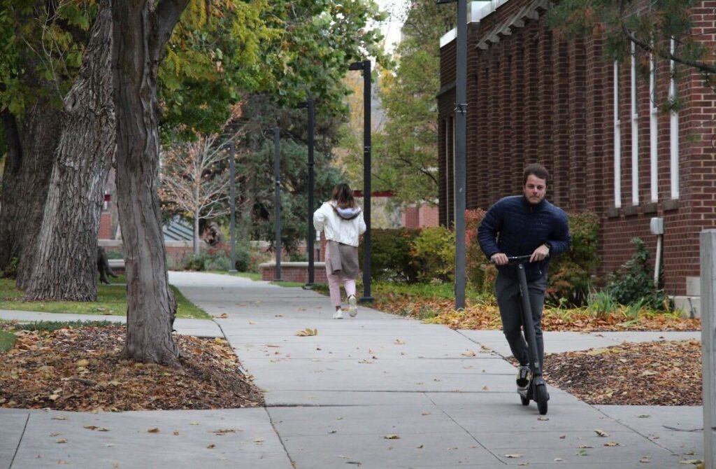 A white man rides an electric scooter through campus with leaves around him.
