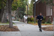 A white man rides an electric scooter through campus with leaves around him.