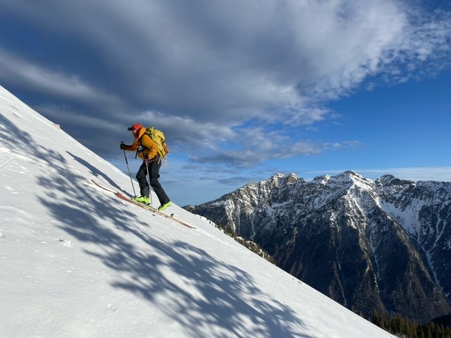 a man hikes up a snowy mountain with skiis and a yellow puffer jacket