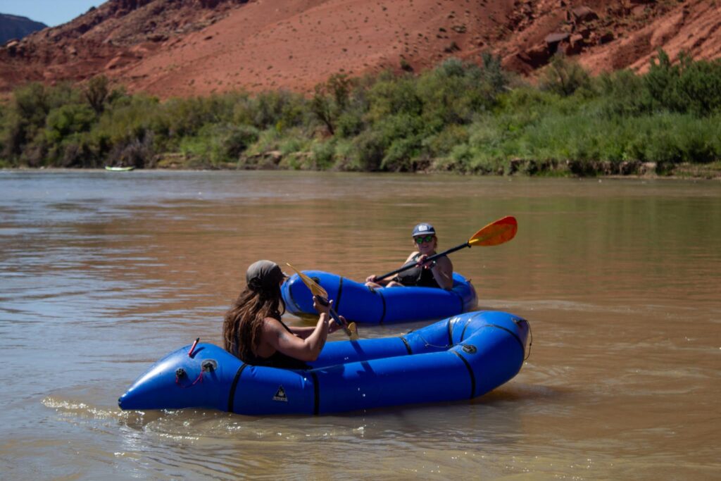Two river rafters sit in blue rafts while holding paddles. They are floating in a river with green foliage on the shore and red rock hills in the background.