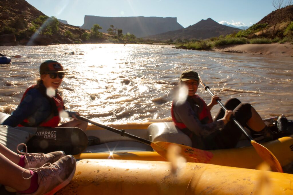 Two river rafters sit in a yellow boat that is on a river in a desert landscape. Mountains can be seen in the background while the sun shines onto the water.